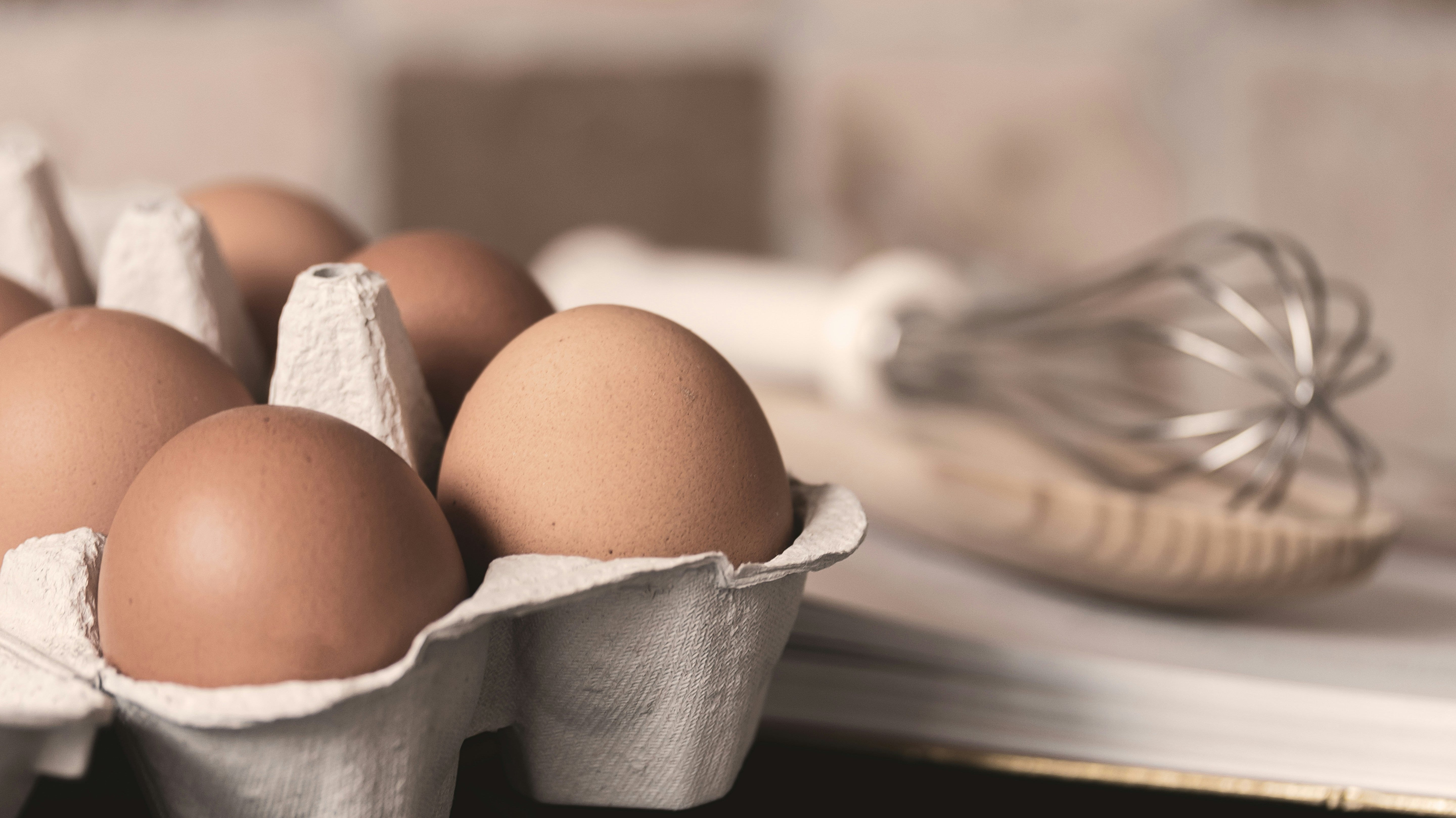 Brown eggs in a carton in a kitchen with a whisk in the background