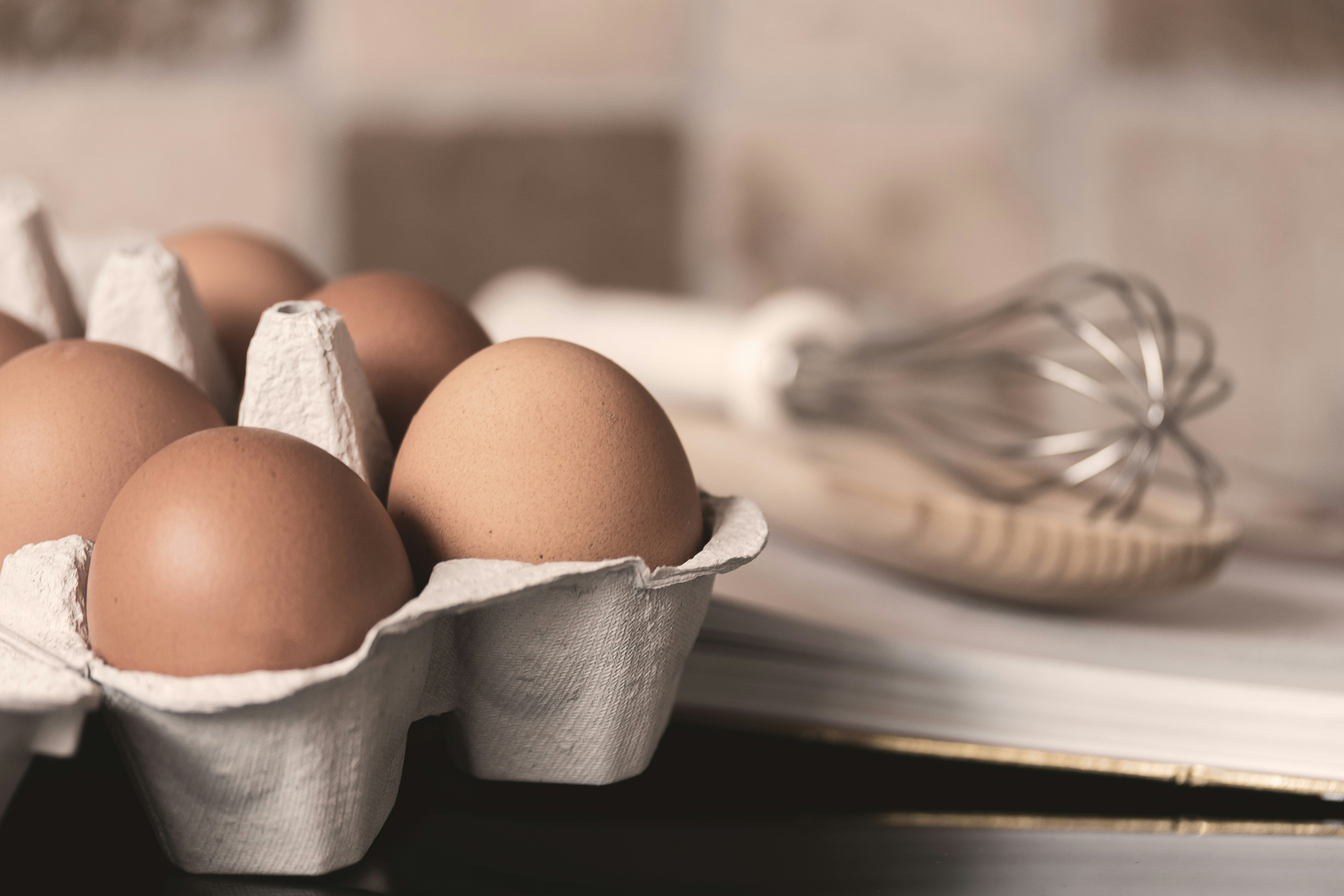 Brown eggs in a carton in a kitchen with a whisk in the background