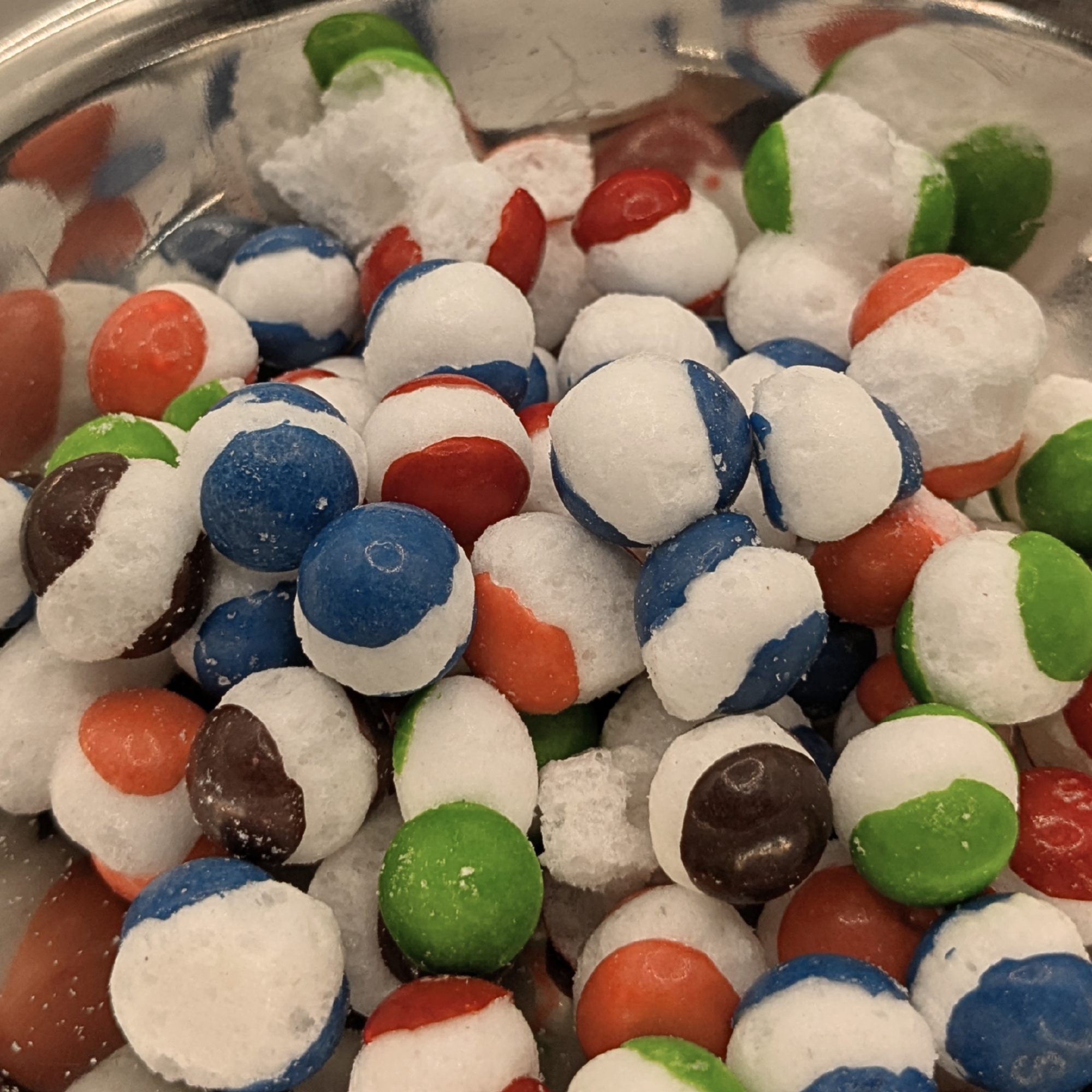A close-up pile of small round candy in a shiny metal bowl. Colors visible include white, blue, red, green, orange,, and purple.