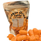 A stand-up pouch of Freeze Dried Taffy Dried Orange snack pieces on a white background, with several orange snack pieces spilled in front. The pouch label reads 'Freeze Dried Taffy Dried Orange' and shows nutrition facts and ingredients.