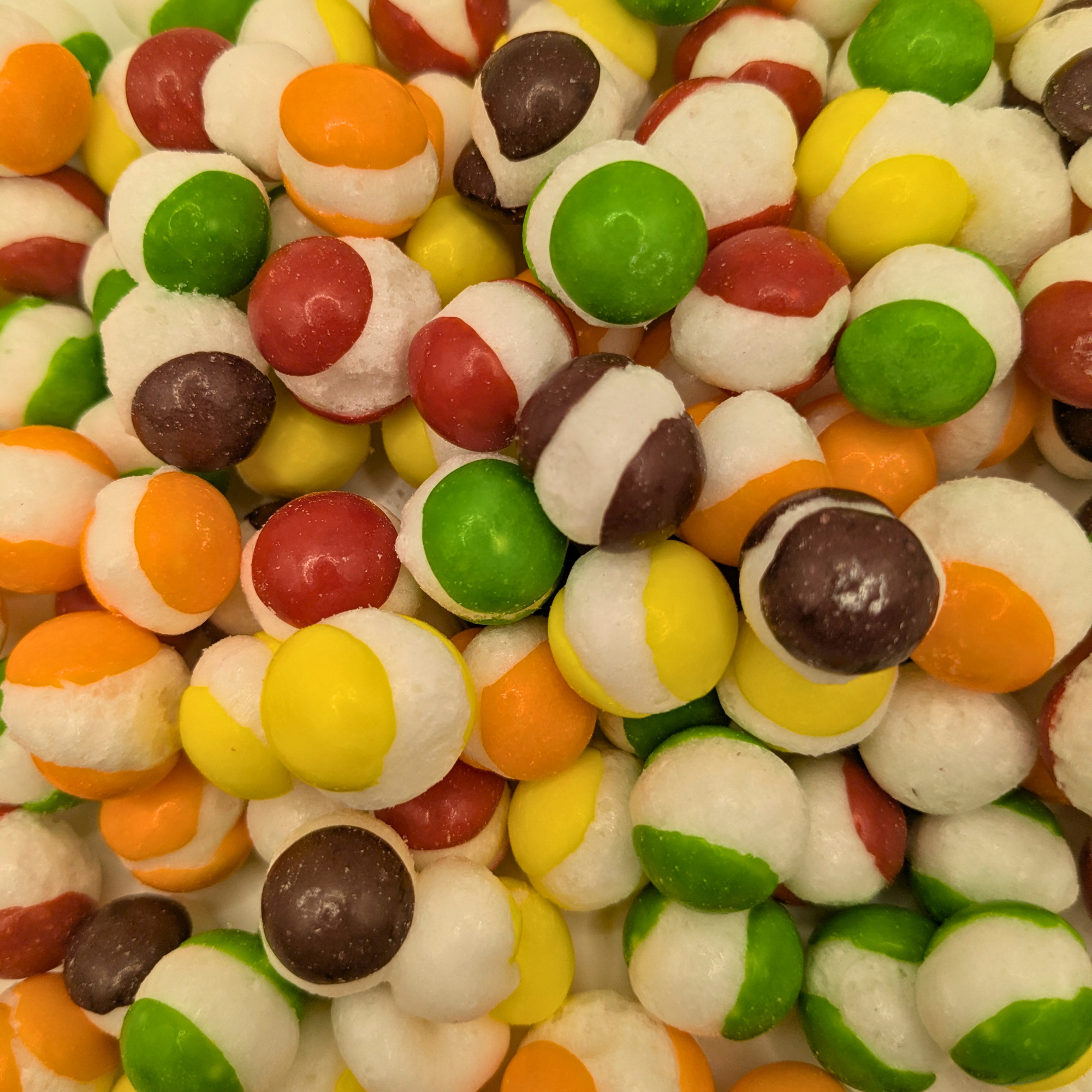 A close-up pile of colorful candy resembling freeze dried Skittles, with round candies in green, yellow, orange, red, and brown with white centers.